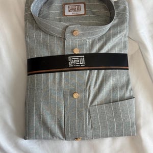 May include: Gray striped shirt with gold buttons and a band collar. The shirt has a pocket and is folded neatly. The text "AL ASEEL" is visible on the label and band.