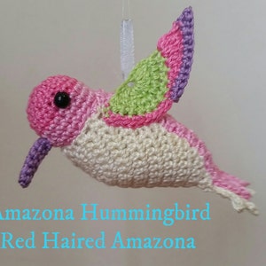 May include: A crocheted hummingbird ornament made with pink, white, green and purple yarn. The hummingbird has a black eye and a long beak. The text "Amazona Hummingbird Red Haired Amazona" is below the hummingbird.