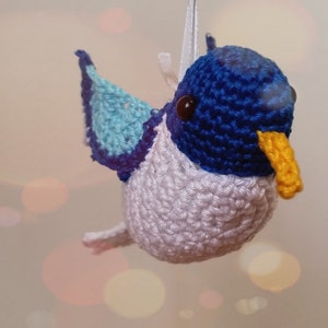 Puede incluir: Un adorno de colibr&iacute; de crochet hecho con hilo azul, blanco y azul claro. El colibr&iacute; tiene un pico amarillo y ojos negros.