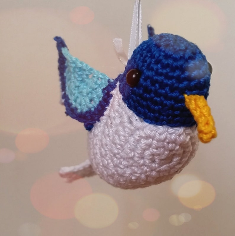Puede incluir: Un adorno de colibr&iacute; de crochet hecho con hilo azul, blanco y azul claro. El colibr&iacute; tiene un pico amarillo y ojos negros.