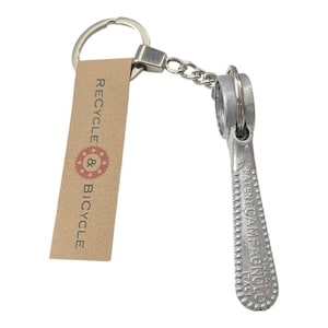 May include: A silver-toned metal keychain featuring a vintage bicycle component shaped like a lever. Attached is a rectangular tan paper tag printed with the text RECYCLE & BICYCLE and a red gear icon. The metal piece is embossed with PATENT CAMPAGNOLO.