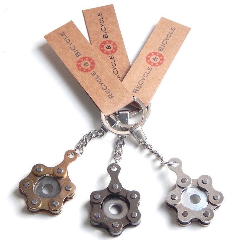May include: Three keychains made from recycled bicycle chains. Each keychain features a star-shaped charm with a silver, gold, or black finish. The keychains are attached to brown paper tags with the text "Recycle & Bicycle" printed on them.