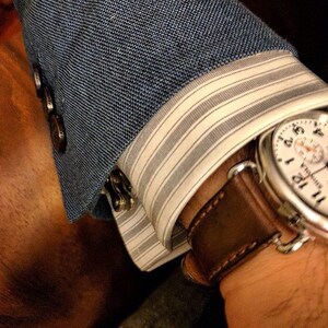 May include: A brown leather watch strap with a white watch face with black markings. The watch is worn on a wrist with a blue blazer and white striped shirt cuff.