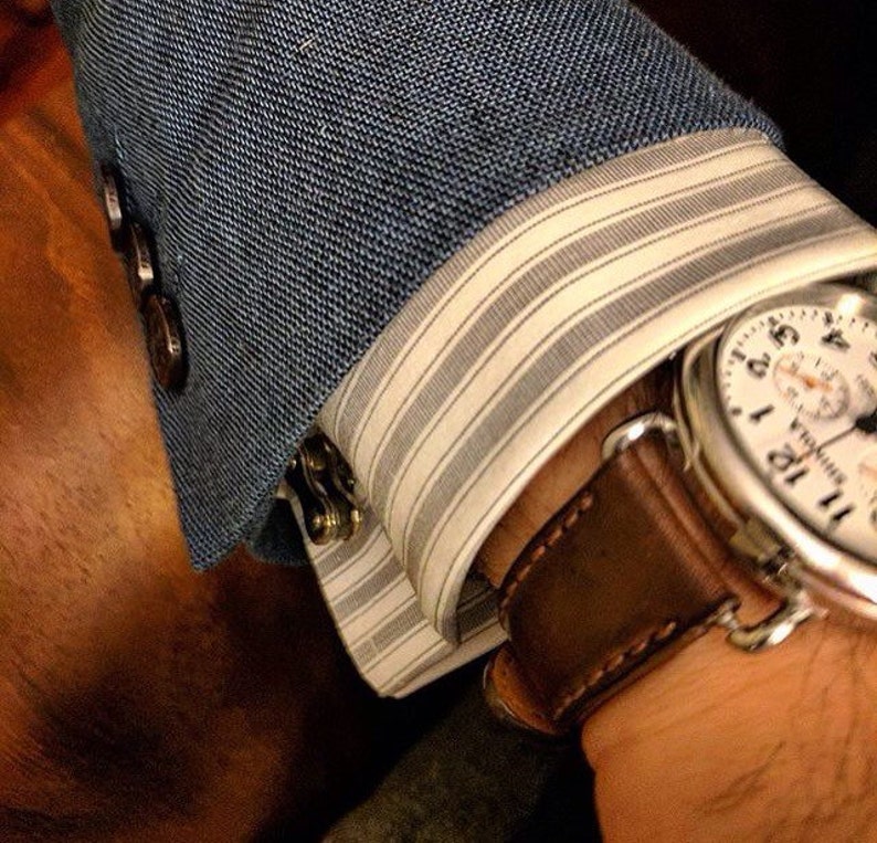 May include: A brown leather watch strap with a white watch face with black markings. The watch is worn on a wrist with a blue blazer and white striped shirt cuff.