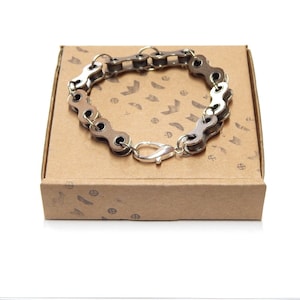 Pode incluir: Um bracelete de corrente de bicicleta prateado com um fecho prateado, apresentado em uma caixa marrom com um padrão repetitivo de flechas e círculos.