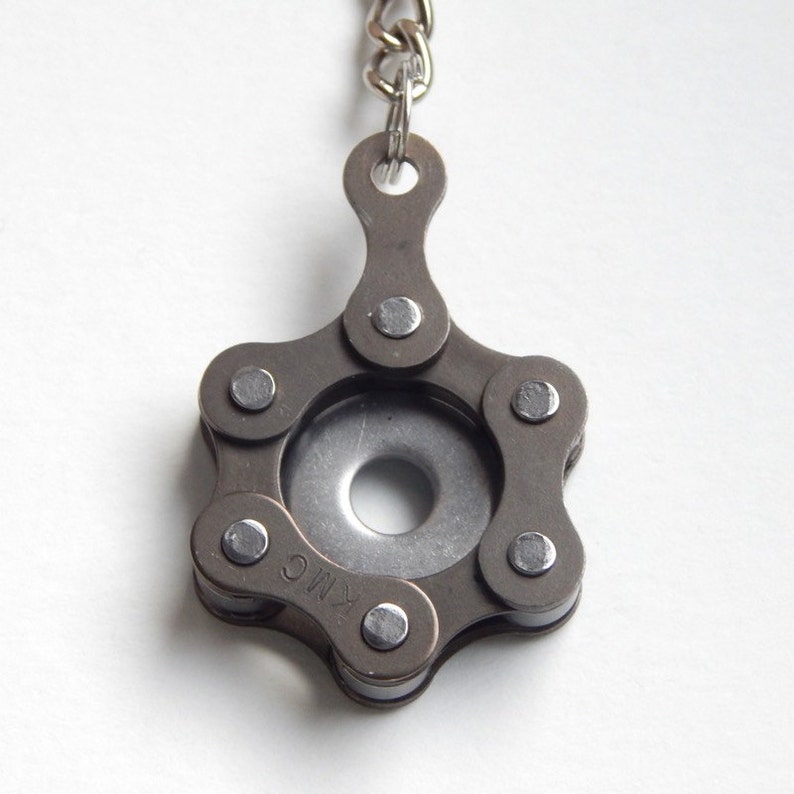 May include: A silver and black metal keychain shaped like a star with a round hole in the center. The keychain is made from a bicycle chain and has the text "KMC" on one of the links.