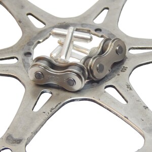May include: Silver cufflinks with a bicycle chain design. The cufflinks are on a silver metal background.