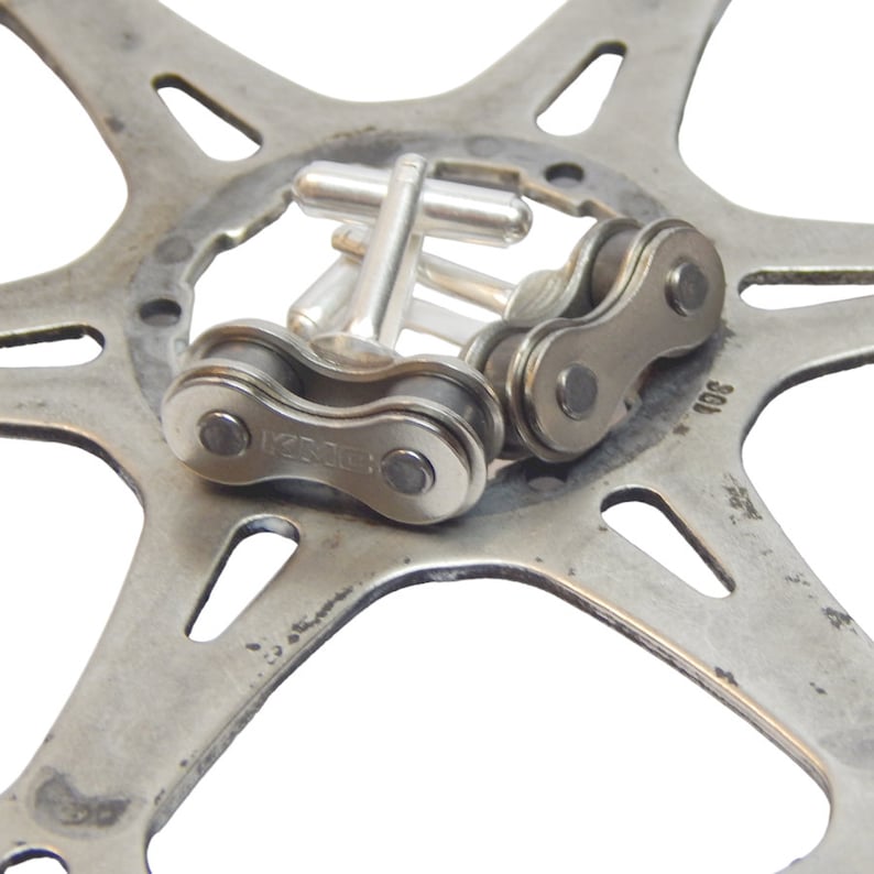 May include: Silver cufflinks with a bicycle chain design. The cufflinks are on a silver metal background.