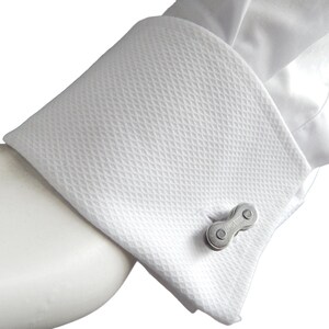 May include: A pair of silver cufflinks shaped like a bicycle chain link. The cufflinks are on a white shirt cuff.