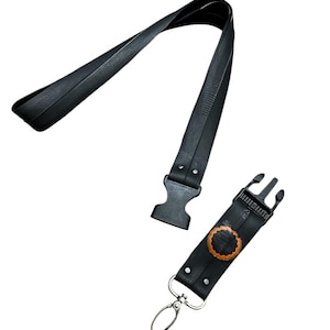 Könnte beinhalten: Schwarzes Leder-Lanyard mit Metallverschluss und Schnalle. Das Lanyard hat ein strukturiertes Design und einen kleinen, befestigten Abschnitt mit einem kreisförmigen, braunen Design. Das Lanyard ist zum Umhängen um den Hals konzipiert.