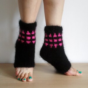 May include: A pair of black knitted leg warmers with a pink heart pattern. The leg warmers are worn on the lower legs and ankles. The background is a light-colored surface.