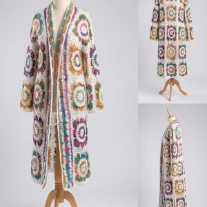 May include: A long, open-front cardigan made of white crochet yarn featuring a repeating pattern of multi-colored floral motifs in shades of pink, yellow, green, and purple. The garment has long sleeves and a straight hem, displayed on a mannequin.