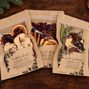 May include: Three kraft paper pouches containing dried fruit and spice simmer pot mixes. Labels read Frosted Orchard, Classic Christmas, and Winter Wonderland. Ingredients include dried apple, orange, cranberry, star anise, and rosemary.