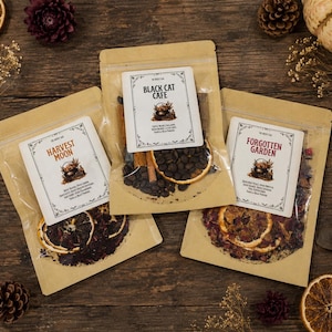 May include: Three kraft paper pouches with clear windows containing dried botanical ingredients. Labels read Harvest Moon, Black Cat Cafe, and Forgotten Garden, each featuring a small cauldron illustration and detailed text descriptions.