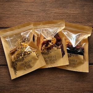 May include: Three stand-up pouches with clear windows containing dried botanical ingredients for simmering. The labels read Orchard Pear, Harvest Orchard, and Bramble Glow. Each pouch includes dried fruit, cinnamon sticks, and spices.