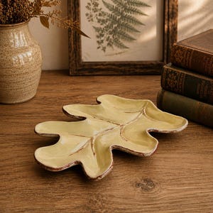 May include: A ceramic dish shaped like an oak leaf with a glossy pale yellow glaze and dark brown distressed edges. The surface features embossed veins that divide the leaf into sections, creating a shallow tray with a smooth, reflective finish.