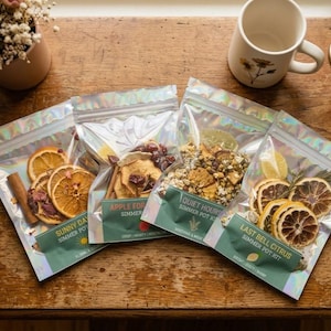 May include: Four clear, iridescent pouches of simmer pot kits on a wooden surface. Each pouch contains dried fruits and herbs. The pouches are labeled "Sunny Day," "Apple For," "Quiet Hours," and "Last Bell Citrus." A mug and a small vase of flowers are in the background.