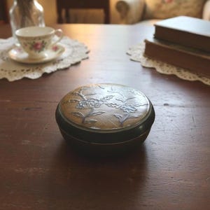 May include: A small, round, antique box with a floral design on the lid. The lid is a light brown color with a darker brown rim. The floral design is in a lighter color, possibly silver or white. The box is sitting on a wooden table.