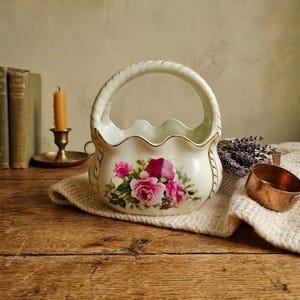 May include: A white ceramic basket with a handle, scalloped rim, and gold trim. The basket is decorated with a floral design of pink and red roses. A vintage-style decorative piece.