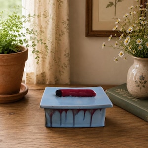 May include: A square ceramic box with a light blue glaze and a dark red, elongated handle on the lid. The sides of the box feature a decorative drip pattern where the dark red glaze runs down over the light blue base, creating a textured effect.
