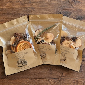 May include: Three brown kraft paper pouches with clear windows containing dried fruit, spices, and botanicals. Each pouch features a label with the text The Mossy Oak, identifying the contents as Orange Spice Cake, Blueberry Leaf, and Grandma's Apple Pie.