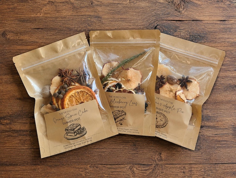 K&ouml;nnte beinhalten: Drei Standbeutel aus Kraftpapier mit getrockneten botanischen Zutaten f&uuml;r einen Simmer Pot. Die Etiketten tragen die Aufschriften The Mossy Oak, Orange Spice Cake, Blueberry Leaf und Grandma's Apple Pie mit Abbildungen von Kuchen, Beeren und Torte.
