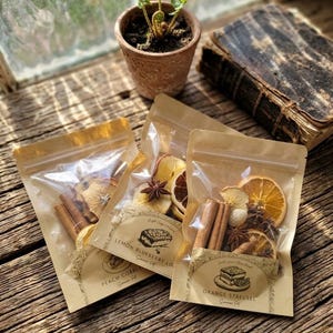 May include: Three clear pouches filled with dried fruit, cinnamon sticks, and star anise, labeled "Peach Cobbler," "Lemon Blueberry Loaf," and "Orange Streusel." A small potted fern and a vintage book are in the background, all on a rustic wooden surface.