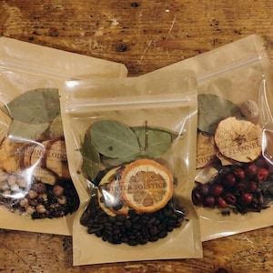 May include: Three clear plastic bags filled with dried botanicals and spices. Each bag contains different ingredients, including dried orange slices, bay leaves, and coffee beans. The bags are sealed and arranged on a wooden surface.