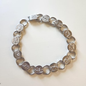 Può includere: Bracciale argentato con maglie circolari con disegni in rilievo. Le maglie sono collegate da piccoli anelli rotondi e ha una chiusura. Il design sulle maglie circolari è un motivo a rilievo.