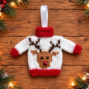 Op de afbeelding: Een miniatuur gebreid trui-ornament met een witte basis en rode boorden bij de kraag, mouwen en onderkant. Het midden toont een bruin rendiergezicht met donkere ogen en een rode neus, afgewerkt met een wit lintje om op te hangen.