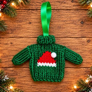 Op de afbeelding: Een miniatuur ornament in de vorm van een groene gebreide trui met een rood-witte kerstmuts afbeelding op de voorzijde. De trui heeft een geribbelde col en een felgroene satijnen lintlus aan de bovenkant om het ornament op te hangen.