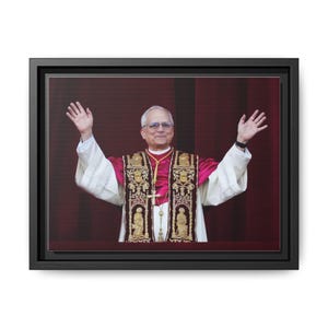 May include: A framed canvas print featuring a man in religious vestments, raising both hands. The man wears glasses and a gold cross. The vestments are white, pink, and gold, with ornate embroidery. The background is a deep red curtain.