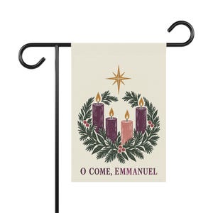 May include: A rectangular garden flag with a cream background featuring an illustration of an Advent wreath with four lit candles, a gold star above, and the text O COME, EMMANUEL in purple at the bottom, suspended from a black metal stand.