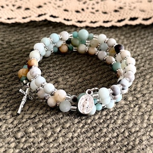 May include: A multi-strand wrap bracelet featuring round beads in shades of pale blue, white, and tan, accented with small silver-toned spacers. The bracelet includes a silver-toned crucifix charm and a small oval medal depicting a religious figure.