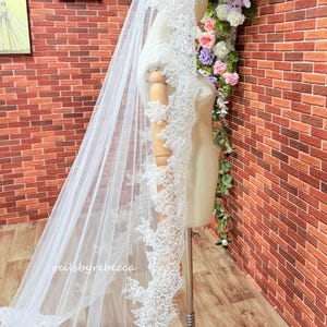 May include: A long, white bridal veil with detailed floral lace edging. The veil is displayed on a mannequin, highlighting its delicate design and flowing fabric. The text "veilsbyrebecca" is visible.