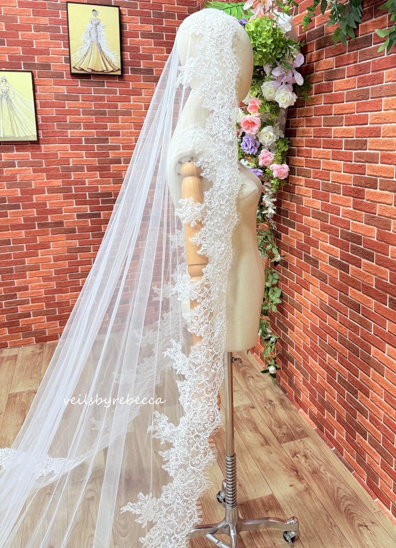 May include: A long, white bridal veil with detailed floral lace edging. The veil is displayed on a mannequin, highlighting its delicate design and flowing fabric. The text "veilsbyrebecca" is visible.