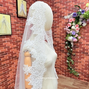 May include: A white bridal veil with detailed lace edging. The veil is displayed on a mannequin, revealing the delicate floral patterns. The background includes a brick wall and floral decorations, emphasising the veil's elegance and design.