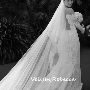 May include: A bride wearing a white lace wedding dress with a long, flowing veil. The veil is sheer and has a delicate pattern. The bride is walking away from the camera, and her back is to the viewer. The text "Veils by Rebecca" is visible on the veil.