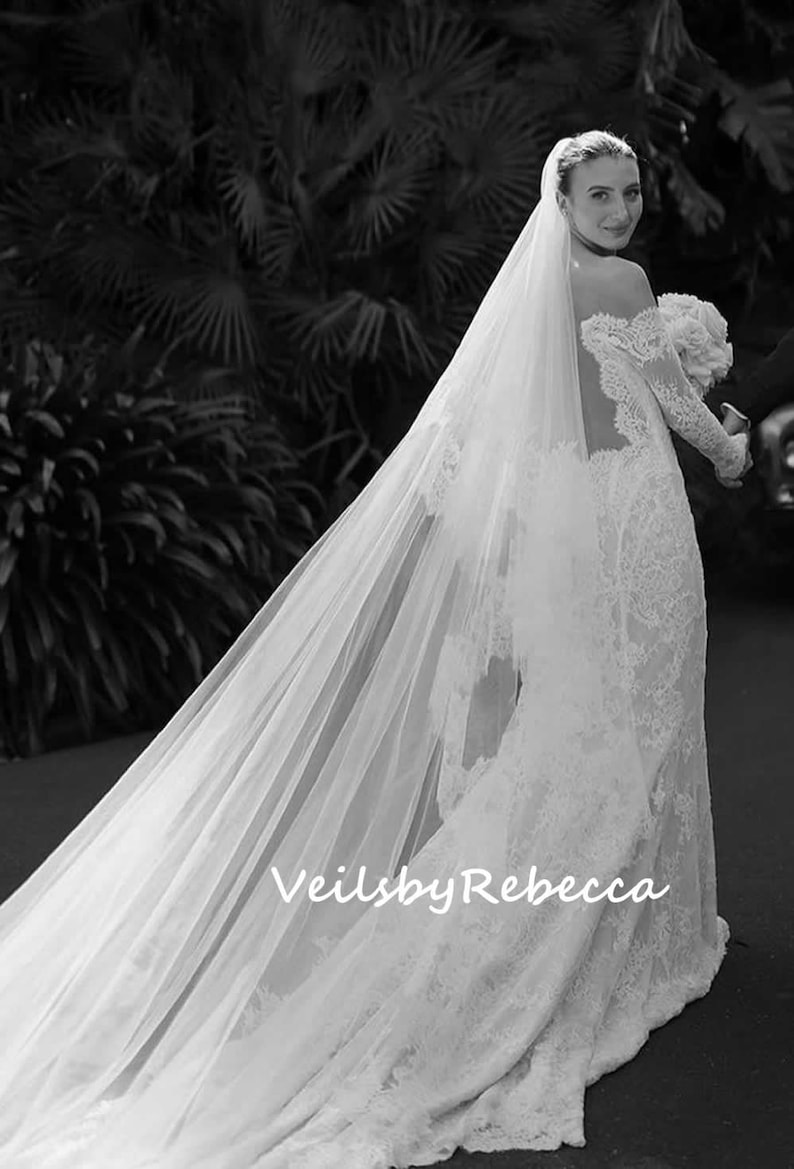 May include: A bride wearing a white lace wedding dress with a long, flowing veil. The veil is sheer and has a delicate pattern. The bride is walking away from the camera, and her back is to the viewer. The text "Veils by Rebecca" is visible on the veil.