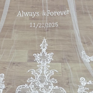 May include: A sheer white wedding veil with delicate lace trim and the words "Always & Forever" and the date "11/22/2025" embroidered in white. The veil is spread out on a light wood surface.