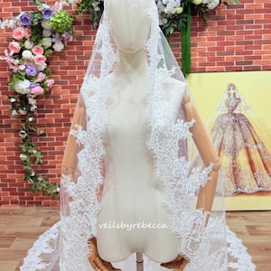 May include: A white bridal veil with detailed lace edging, shown on a mannequin. The veil has delicate floral patterns and is made of sheer material. The text "veilsbyrebecca" is visible on the veil.