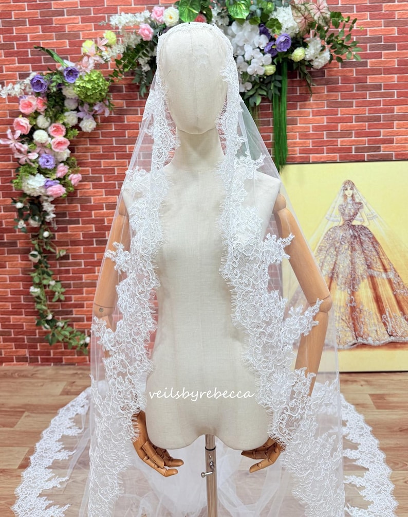May include: A white bridal veil with detailed lace edging, shown on a mannequin. The veil has delicate floral patterns and is made of sheer material. The text "veilsbyrebecca" is visible on the veil.