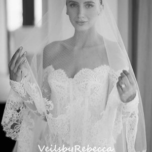 May include: A bride wearing a white lace wedding dress with a long tulle veil. The veil is draped over her head and shoulders, and she is looking at the camera. The text "VeilsbyRebecca" is visible in the lower center of the image.