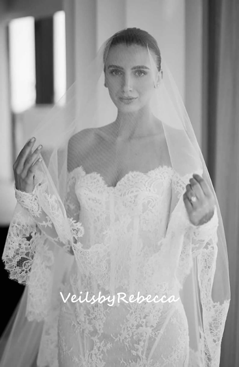 May include: A bride wearing a white lace wedding dress with a long tulle veil. The veil is draped over her head and shoulders, and she is looking at the camera. The text "VeilsbyRebecca" is visible in the lower center of the image.