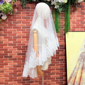 May include: A white bridal veil draped over a mannequin, featuring intricate floral lace embroidery along the scalloped edges. The sheer tulle fabric extends downward, showcasing detailed needlework patterns and the text veilsbyrebecca.