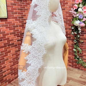 May include: A white lace bridal veil with detailed floral patterns. The veil is displayed on a mannequin, highlighting its delicate design. The text "veilsbyrebecca" is visible on the mannequin. The background includes a brick wall and floral arrangements.