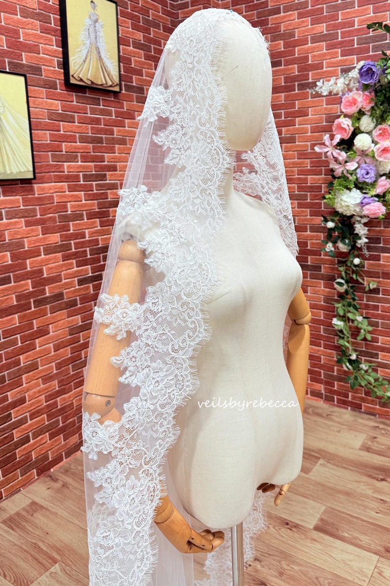 May include: A white lace bridal veil with detailed floral patterns. The veil is displayed on a mannequin, highlighting its delicate design. The text "veilsbyrebecca" is visible on the mannequin. The background includes a brick wall and floral arrangements.