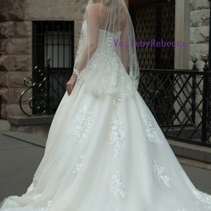 May include: A strapless ivory wedding gown with floral lace appliques and a flowing skirt. The bride is wearing a sheer veil with delicate lace trim. The text "VeilsbyRebecca" is visible on the image.