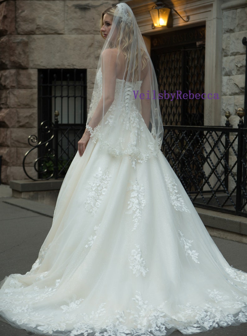 May include: A strapless ivory wedding gown with floral lace appliques and a flowing skirt. The bride is wearing a sheer veil with delicate lace trim. The text "VeilsbyRebecca" is visible on the image.