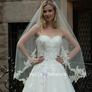 May include: A white bridal veil with a delicate lace trim. The veil is made of soft tulle and has a single layer. The lace trim is a floral design with a scalloped edge.